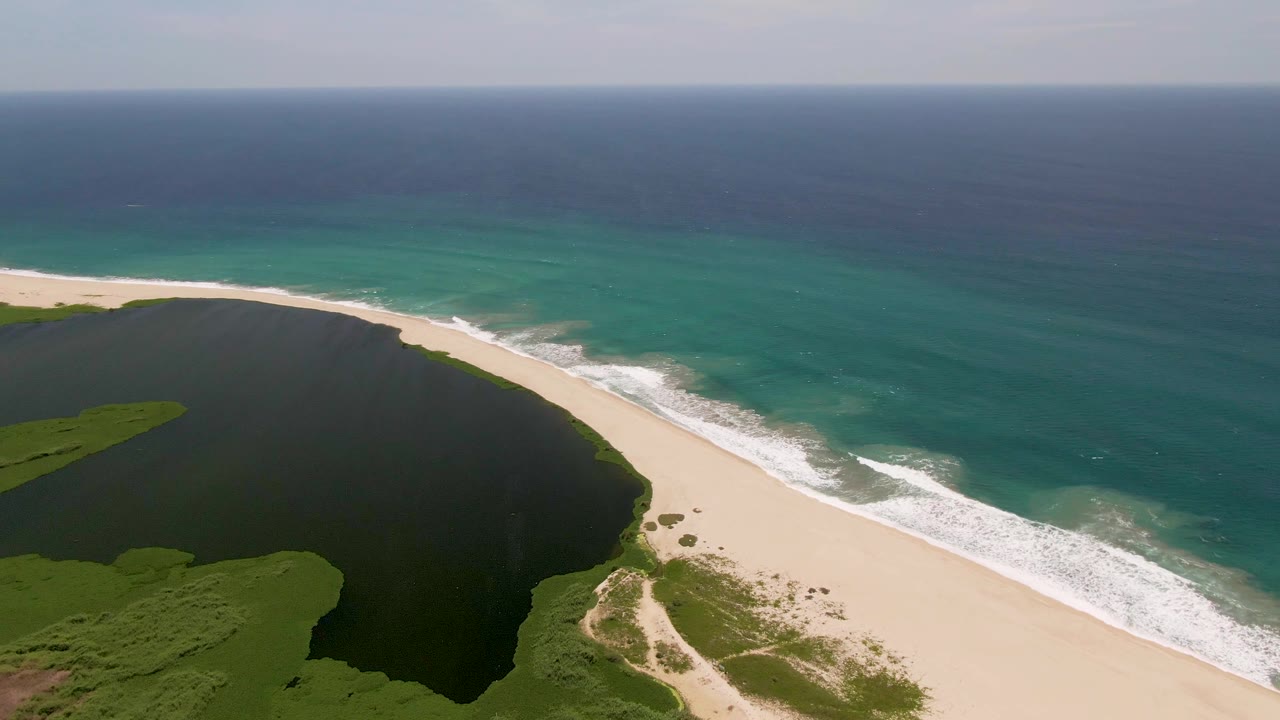 San Jose Del Cabo aerial view with dark lagoon water on the left, bright sand through the center, layered turquoise ocean on the right, scattered surf foam, and no visible people.