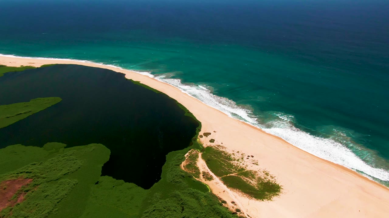 Aerial view of a dark lagoon beside a sandy beach and turquoise ocean.