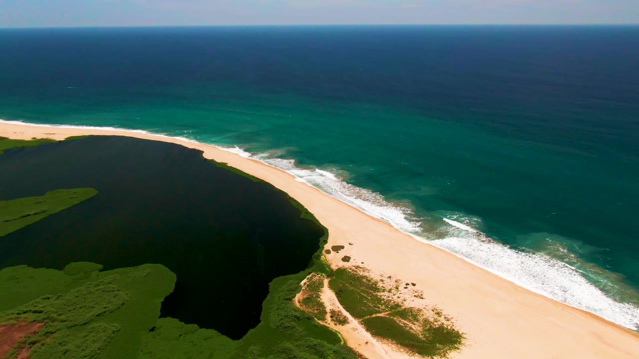 Aerial view over San Jose Del Cabo coastline with white surf advancing along the sandy shore beside the dark lagoon and no visible people.