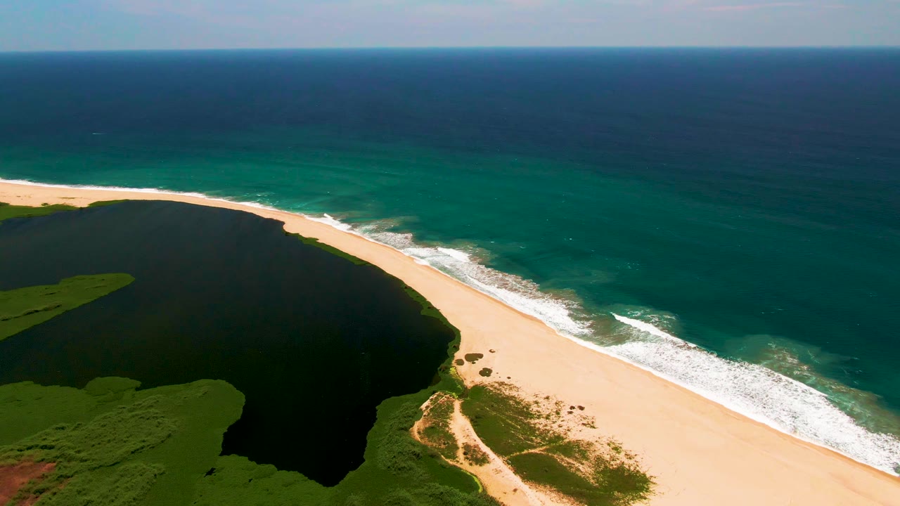 Wide aerial frame in San Jose Del Cabo with more open ocean visible above the beach, the dark lagoon at left, and no visible people on the sand.