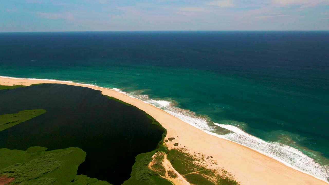 Aerial view over San Jose del Cabo showing a dark green lagoon beside a pale sand beach, white surf, blue-green ocean, and no visible people.