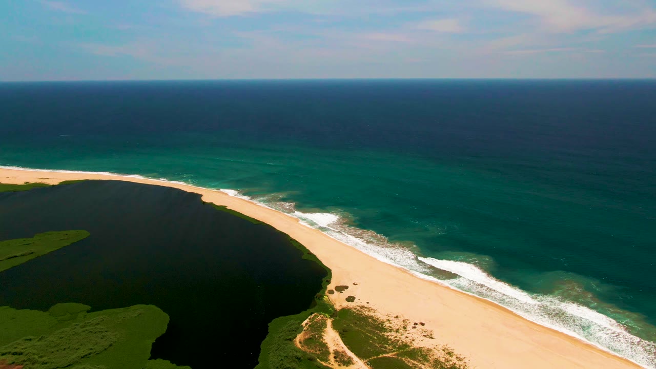 San Jose del Cabo aerial still with dark lagoon water on the left, a curved sandy shoreline in the center, textured surf on the right, and no visible people.