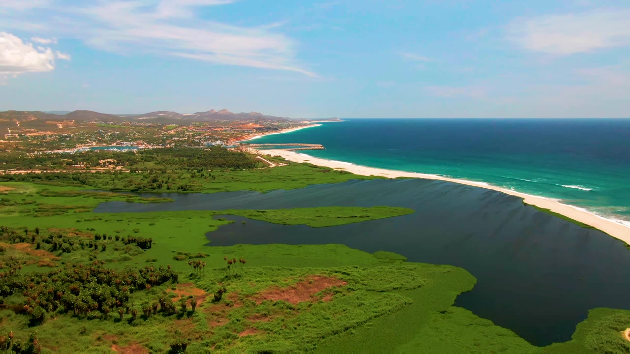 Opening aerial frame over San Jose del Cabo with green wetland fields in front, a dark lagoon, a bright sandbar, turquoise surf, distant hills, and no visible people.