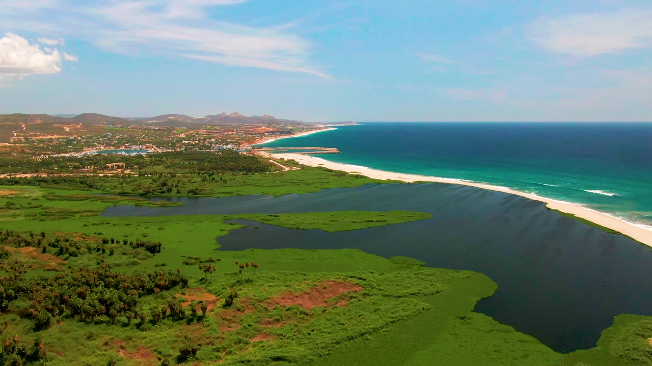 Aerial view in San Jose del Cabo one second in, with the lagoon and green fields filling the lower frame, the sandbar stretching toward the right, ocean surf beyond it, and no visible people.