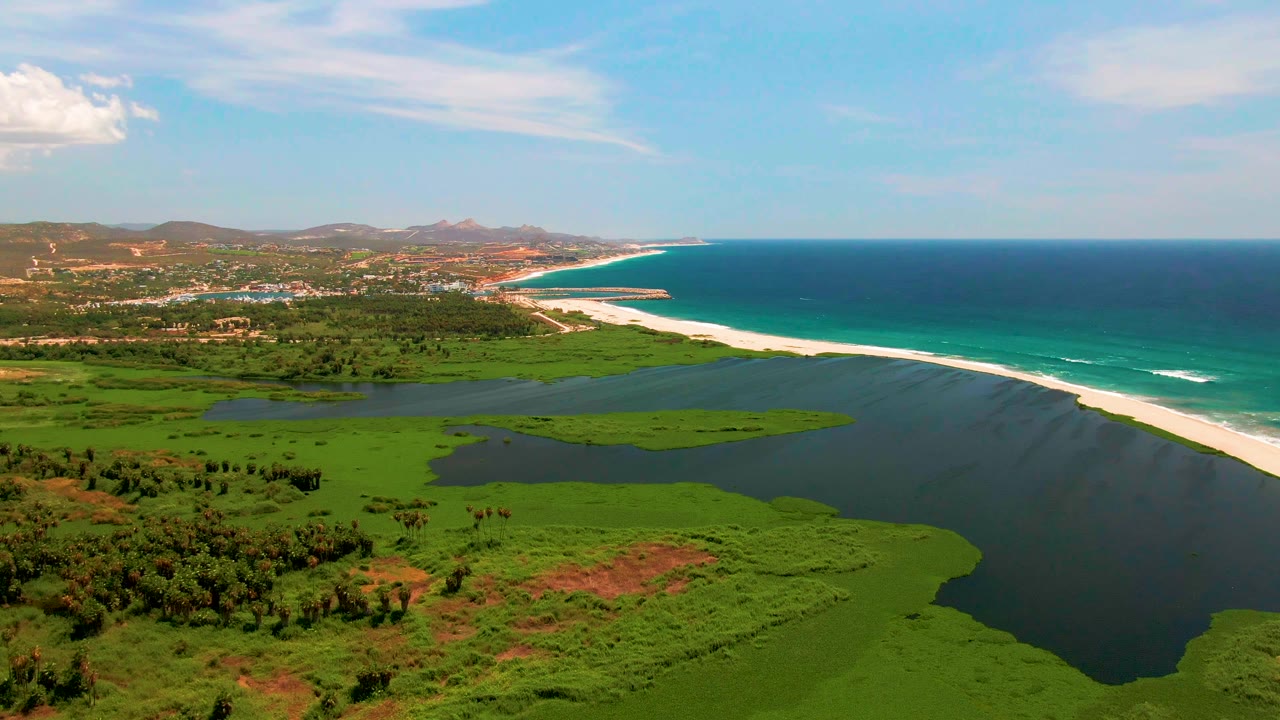 San Jose del Cabo aerial frame showing white ocean waves along the outer beach, darker lagoon water inside the sandbar, green fields below, low hills at left, and no visible people.