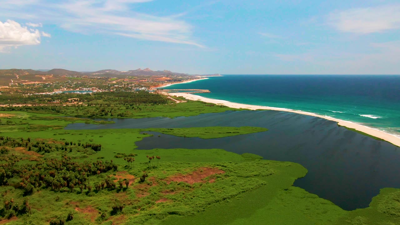 Mid-clip aerial view over San Jose del Cabo with green wetland patches in the foreground, a broad lagoon, a long white beach, blue ocean to the right, distant hills, and no visible people.