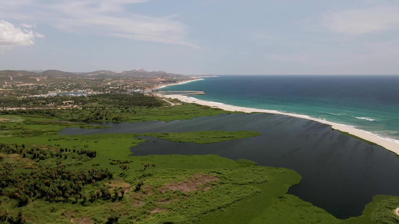 Aerial frame over San Jose del Cabo showing green lagoon vegetation, dark lagoon water, a pale beach, turquoise ocean, distant hills, and no visible people.