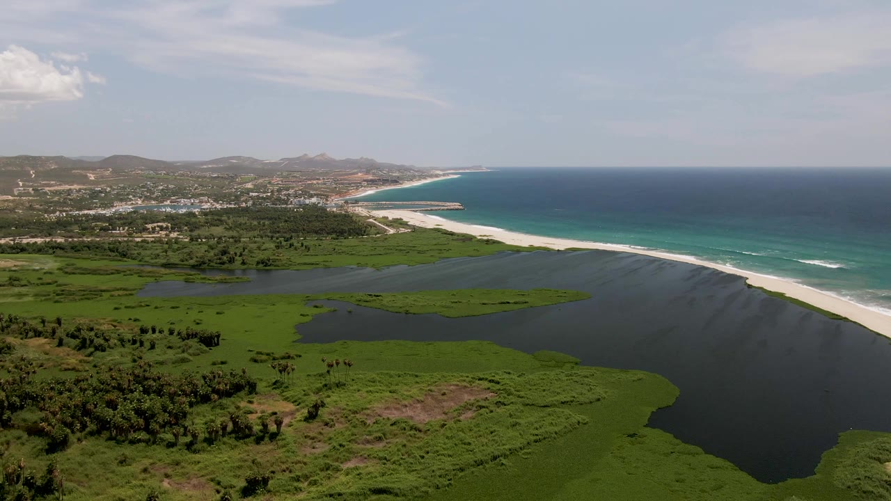 A second aerial frame over San Jose del Cabo holds the lagoon foreground, bright beach edge, blue ocean, distant coastal development, hills, clouds, and no visible people.