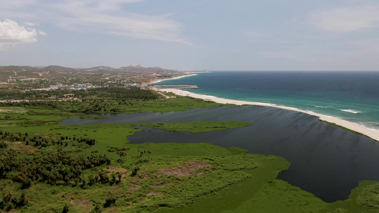 Aerial view in San Jose del Cabo with the lagoon filling the lower frame, the beach and surf running along the right side, distant town structures, hazy hills, clouds, and no visible people.