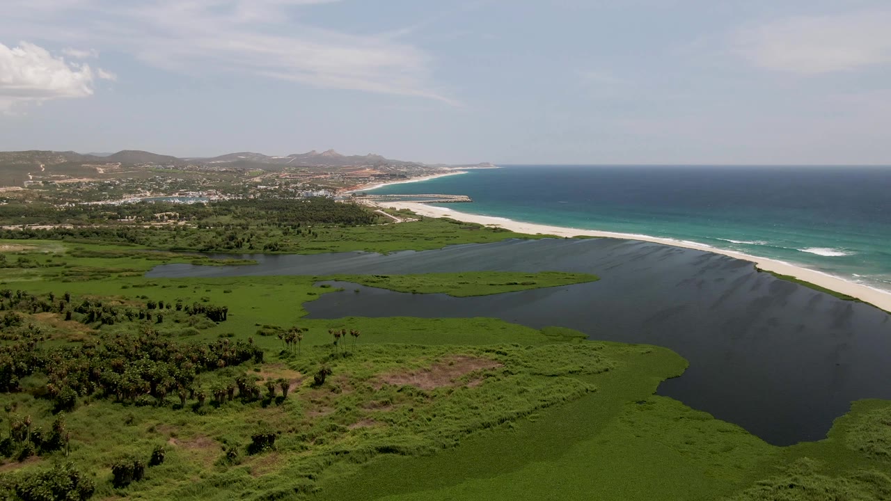 Opening aerial frame over San Jose del Cabo showing green wetland and dark lagoon water beside a pale sand beach and turquoise ocean, with no visible people.