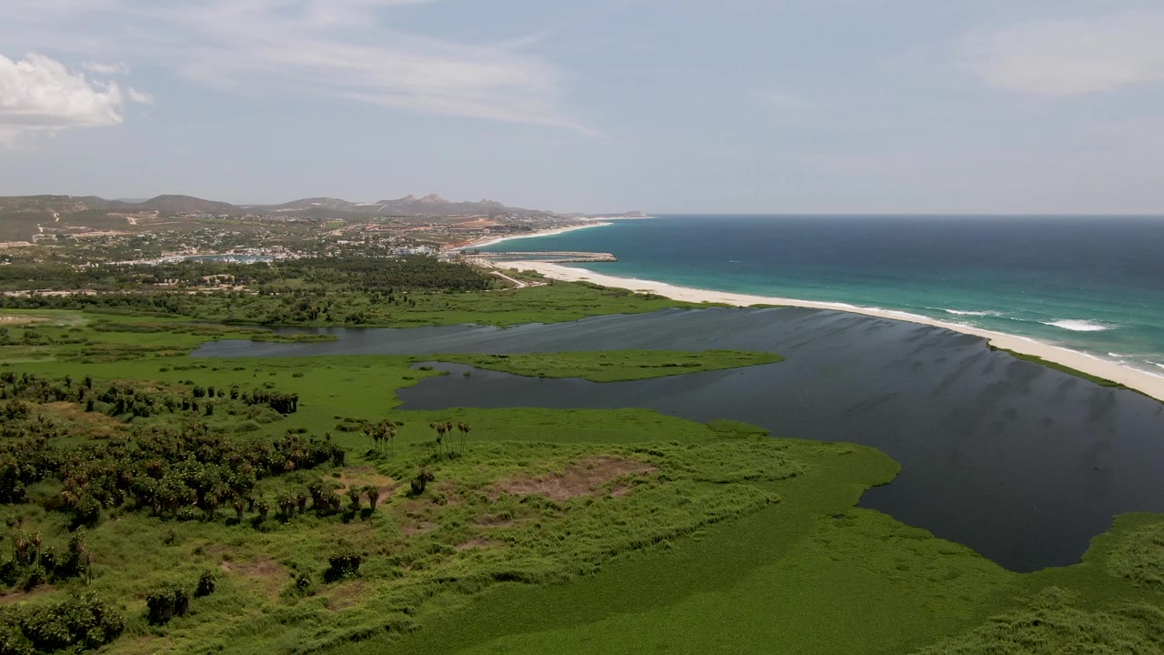 One second into the clip, the San Jose del Cabo lagoon and marsh remain in the foreground while the white beach and ocean surf line stretch toward distant hills, with no visible people.