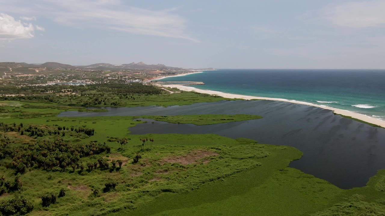 At 0:02, the dark lagoon water contrasts with bright green wetland and turquoise ocean beyond the sandbar in San Jose del Cabo, with no visible people.