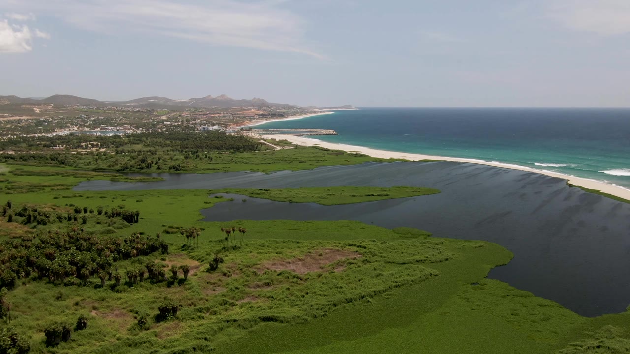 Midway through the clip, San Jose del Cabo appears as a wide coastal panorama with lagoon in the foreground, beach and surf across the center, and hills under a pale sky, with no visible people.