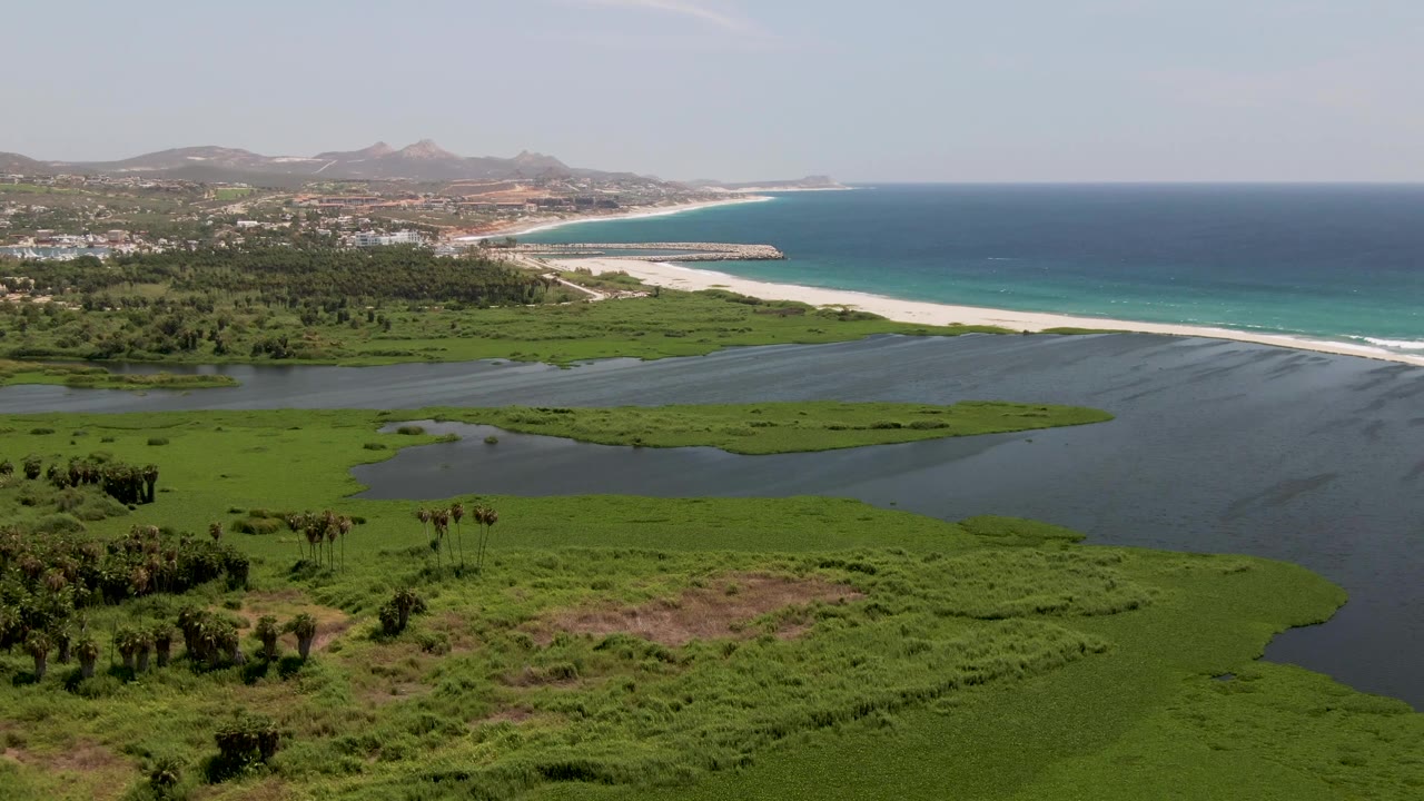 Wide aerial frame in San Jose Del Cabo with marsh vegetation spreading across the foreground, dark lagoon water, bright sandbar and beach, blue ocean, distant buildings, hills, and no visible people.