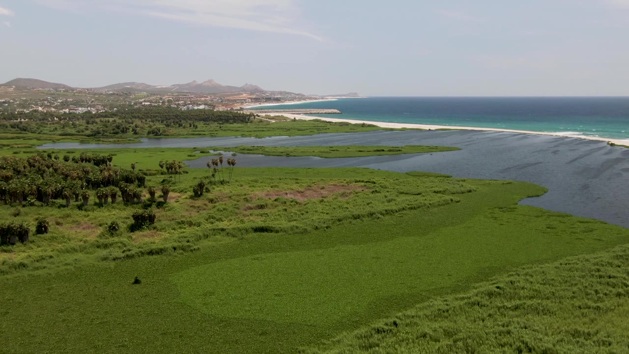 Opening frame over San Jose Del Cabo coastal wetlands with green marsh in front, lagoon water, a pale sand beach, turquoise ocean, distant hills, and no visible people.