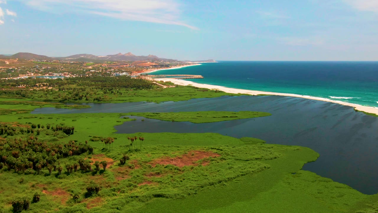 An aerial frame over San Jose del Cabo shows wetland islands in the lagoon, ocean surf running along the beach, built shoreline in the distance, and no visible people.