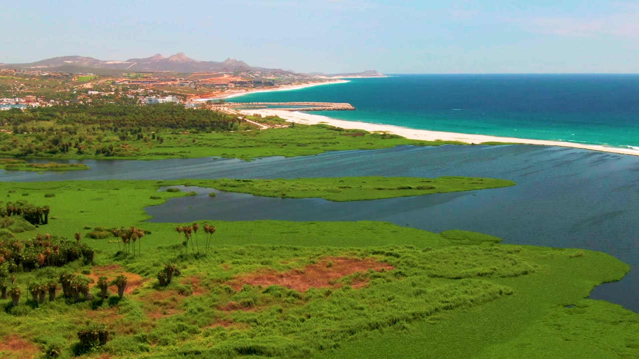 Aerial frame from San Jose del Cabo showing green lagoon vegetation in front, blue water channels, a white beach, turquoise ocean, distant mountains, and no visible people.