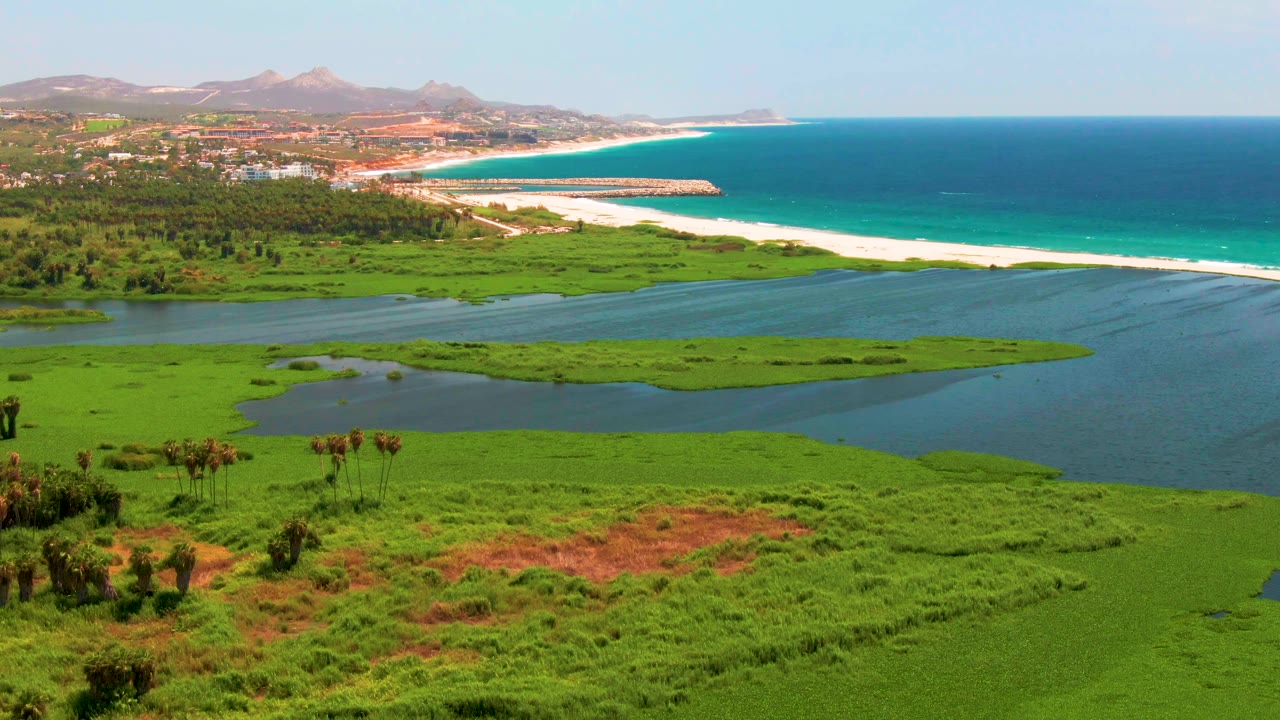 At 0:02, green wetland patches and lagoon water fill the lower frame, with San Jose del Cabo beach, open ocean, distant hills, and no visible people beyond.