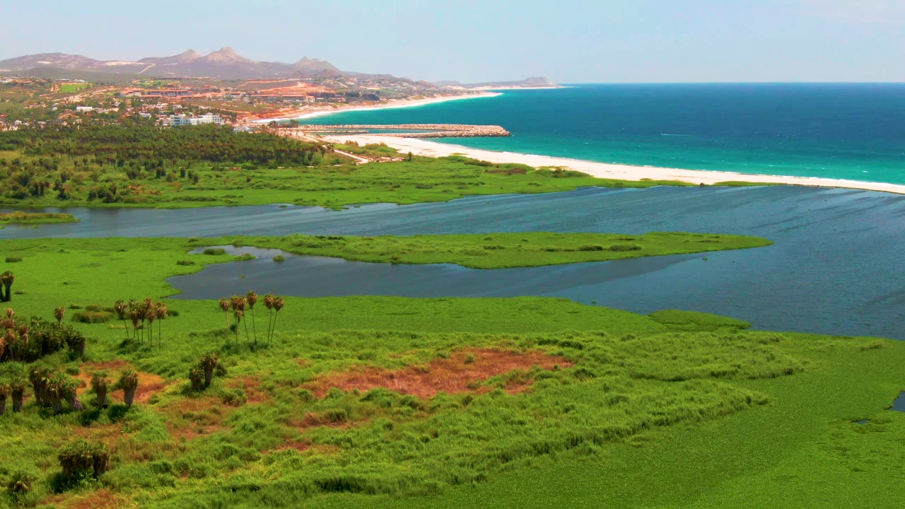 At 0:03, the aerial composition shows lagoon marshland and tidal water below, a long beach and turquoise ocean above, coastal buildings near the shore, and no visible people.