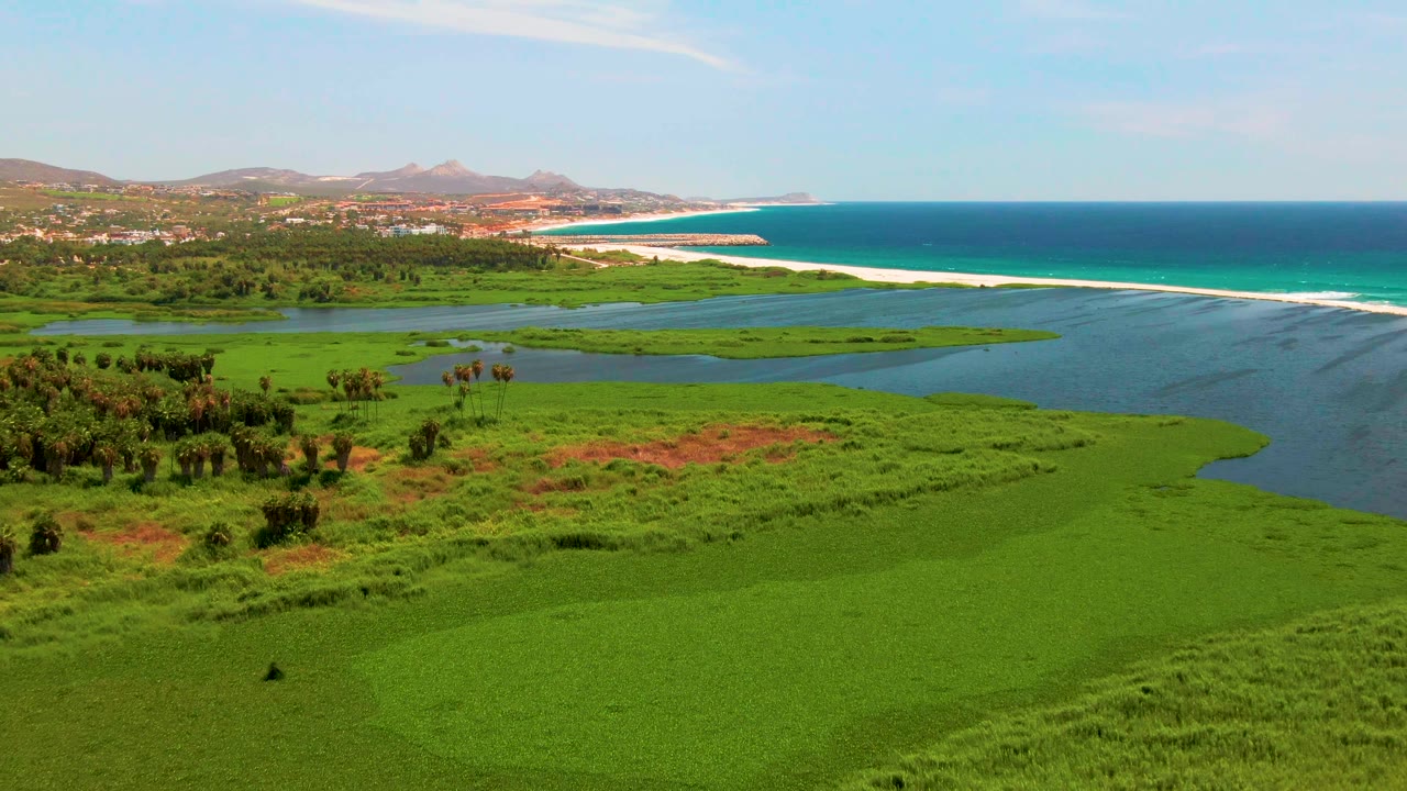 The second aerial frame keeps San Jose Del Cabo wetlands across the foreground, with lagoon channels, a long sand strip, blue ocean, hazy hills, and no visible people.