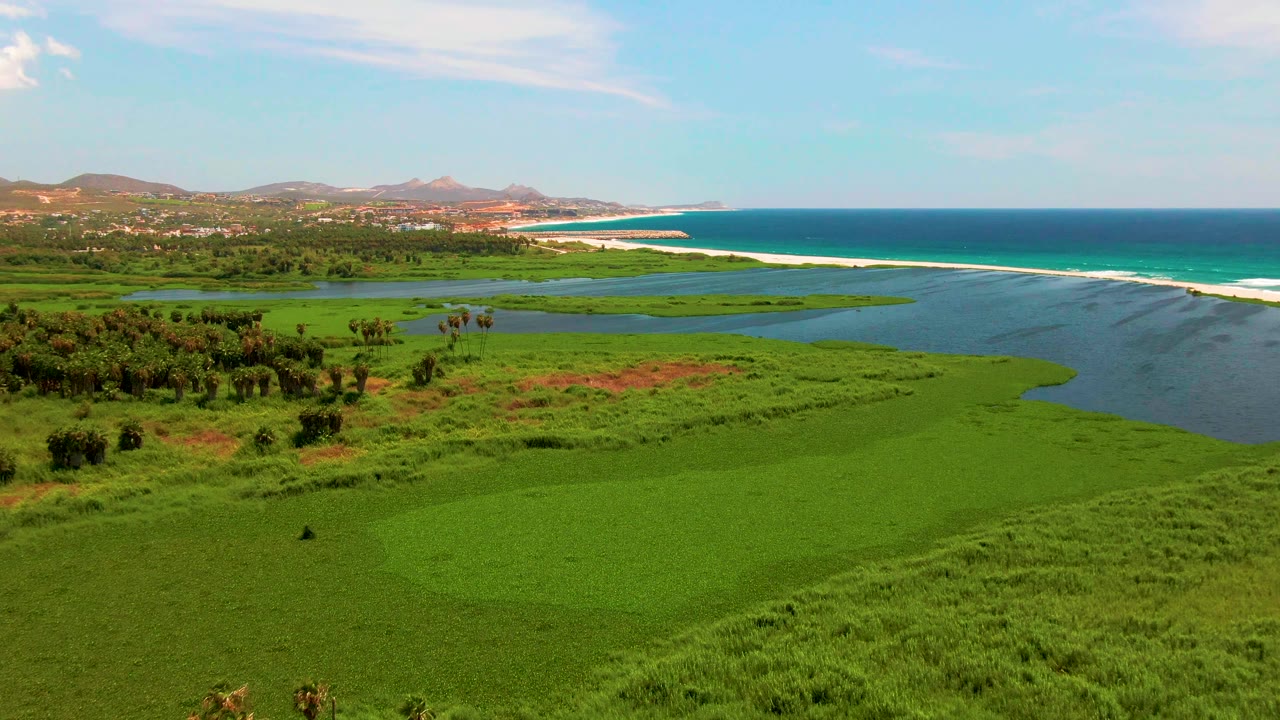 Midway through the clip, San Jose Del Cabo wetlands fill the lower frame while lagoon water, a pale sand beach, turquoise ocean, low mountains, and no visible people remain in view.