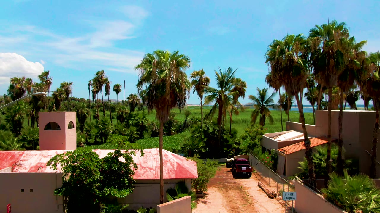 Opening elevated frame in San Jose del Cabo shows beige buildings, a red roof, a dirt path, palms, green fields, distant ocean, blue sky, and no visible people.