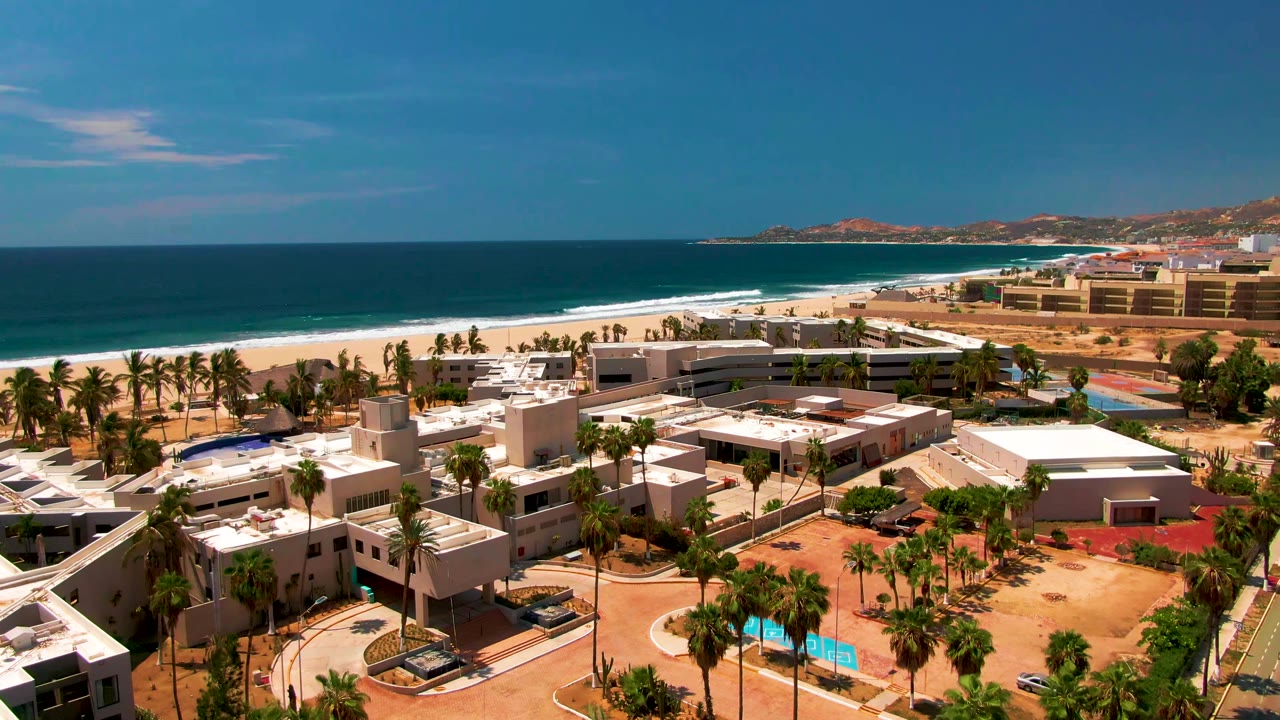 Aerial view over a San Jose del Cabo beachfront resort with white buildings, palm trees, sandy beach, blue ocean, distant coastline, and no visible people.