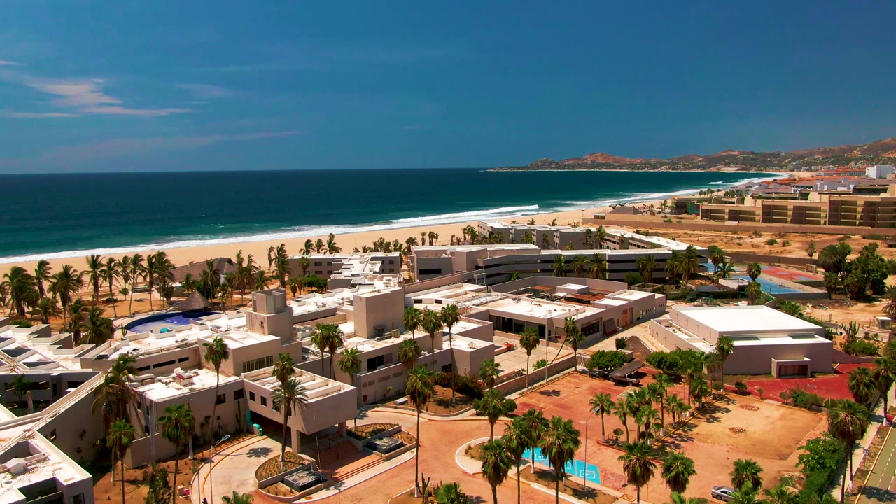 Elevated frame of the San Jose del Cabo resort area with flat white roofs in the foreground, palms and sand in the middle distance, ocean beyond, and no visible people.