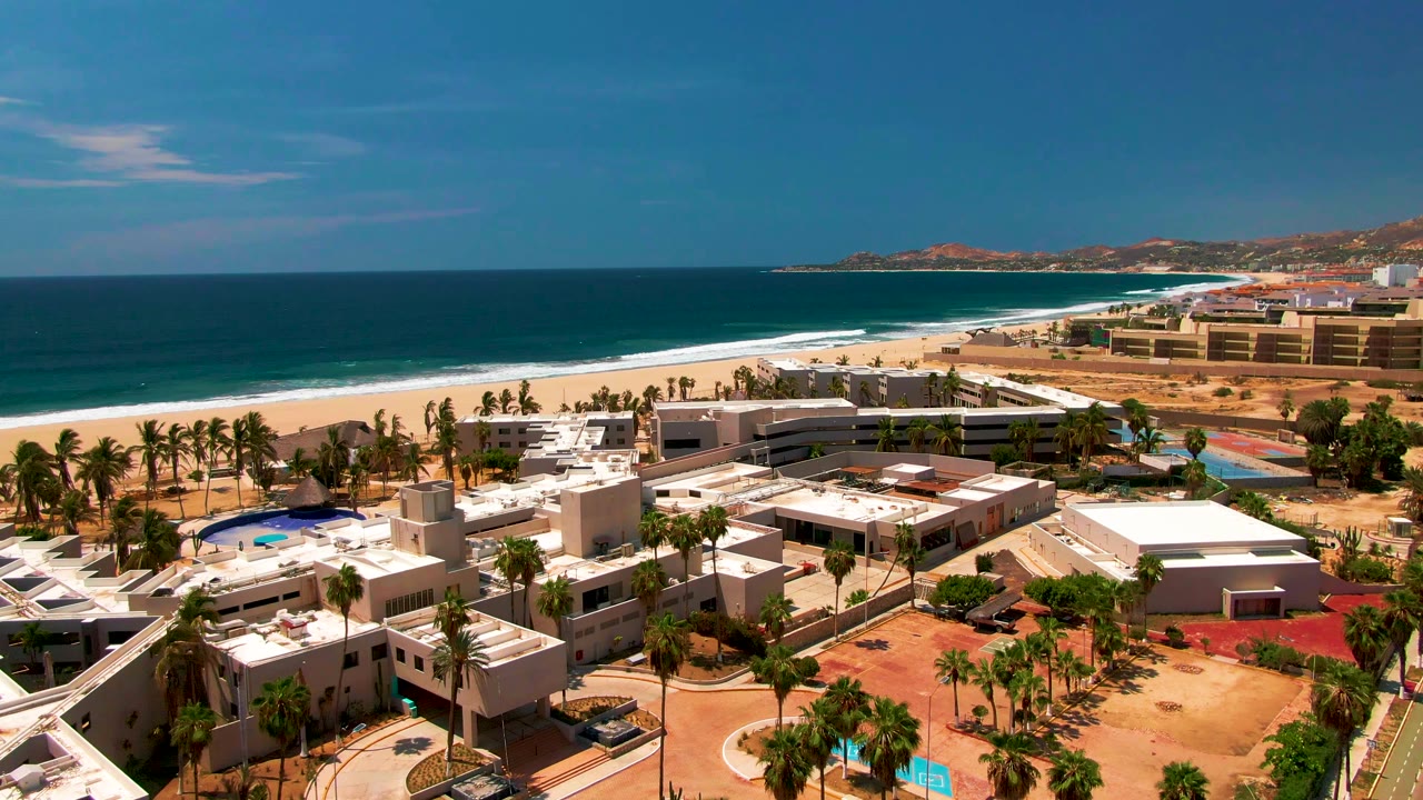 Wide San Jose del Cabo aerial frame showing palm-lined resort grounds, low white buildings, a sandy shore, dark blue water, distant coastal terrain, and no visible people.