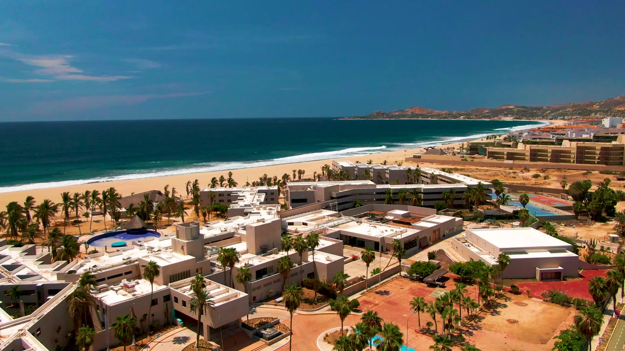 Aerial coastal resort scene with pool, palms, beach, and ocean horizon.