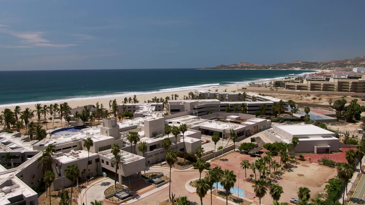 High oblique view in San Jose Del Cabo showing white resort structures, a road through the property, palms, beach, ocean, and a long horizon, with no visible people.