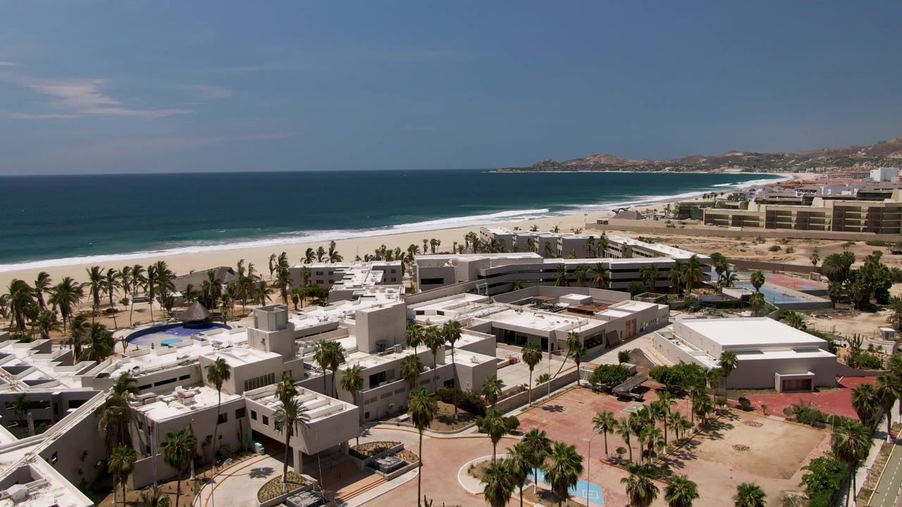 Aerial frame from San Jose Del Cabo with resort rooftops and palms in the lower frame, a curved road near the buildings, white beach, blue water, and no visible people.
