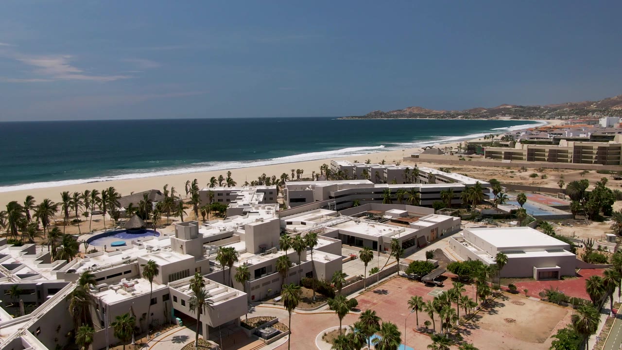 Mid-clip aerial over San Jose Del Cabo with white resort buildings at the bottom edge, palm rows, sandy beachfront, blue ocean, and distant coastal hills, with no visible people.