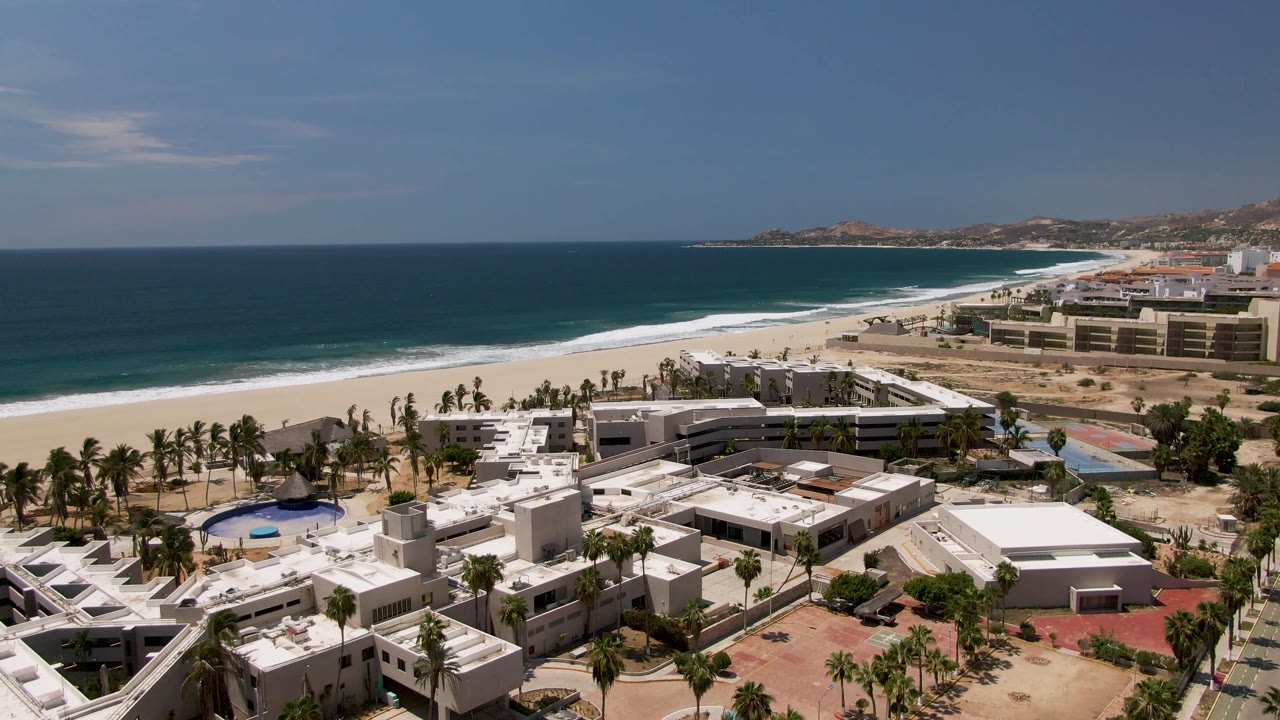 Aerial view over a San Jose Del Cabo beachfront resort with white roofs, palm trees, a sandy shoreline, blue ocean, distant hills, and no visible people.