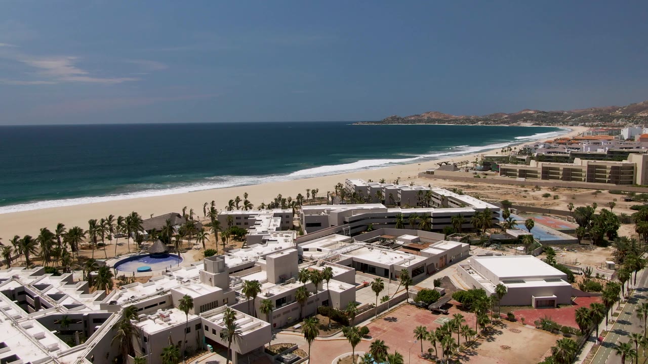 High aerial frame of San Jose Del Cabo resort rooftops and palms beside a broad sandy beach, with surf bands moving along the blue water and no visible people.