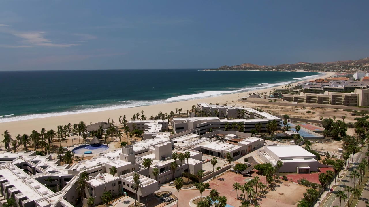 Drone view in San Jose Del Cabo showing the resort grounds slightly shifted across frame, with palms, beach, white surf, blue ocean, far hills, and no visible people.