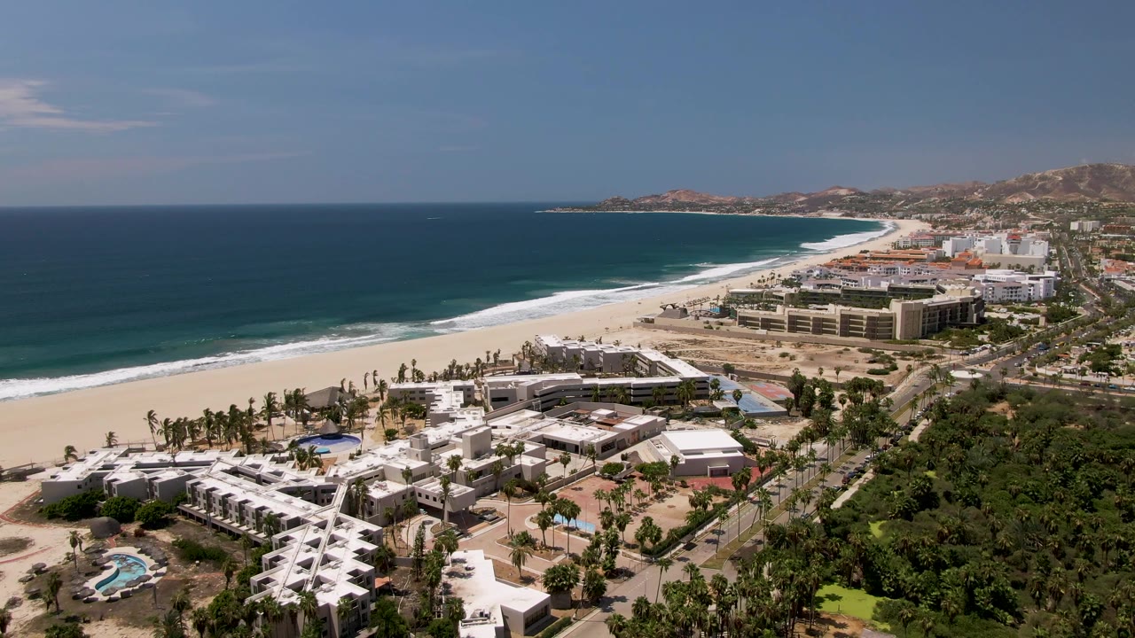 Aerial view of San Jose Del Cabo coastline with blue ocean, white sand beach, beachfront buildings, palm trees, green hillside, and no visible people at the start of the clip.