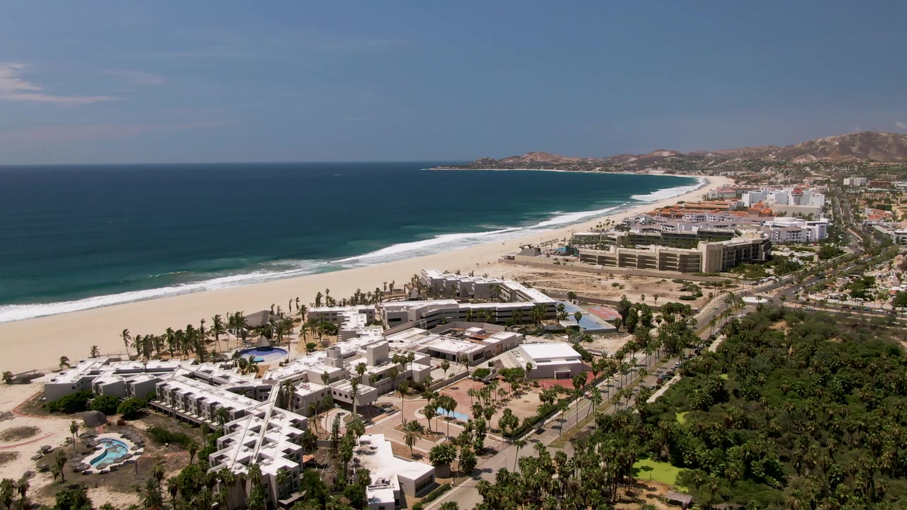 San Jose Del Cabo aerial frame with the surf line extending along the white beach, resort-scale buildings near the shore, roadway traffic, foreground palms, and no visible people.