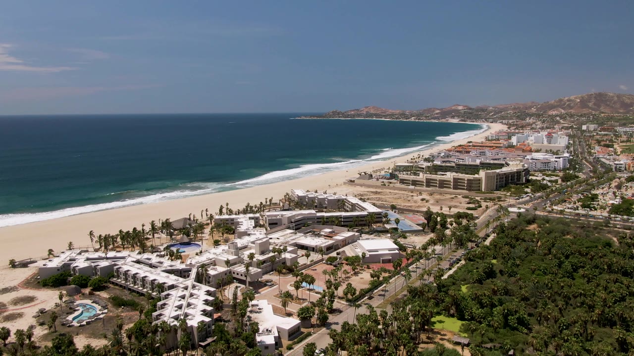 Wide San Jose Del Cabo aerial view with more foreground greenery on the right, a curved beach and surf line across the center, beachfront development, road traffic, and no visible people.