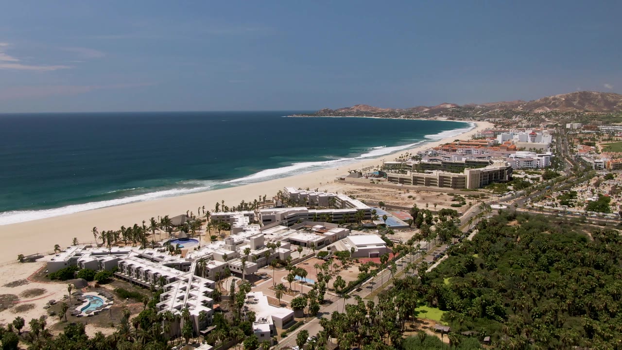 Aerial beach scene with foreground buildings, palms, coastal road, and ocean.