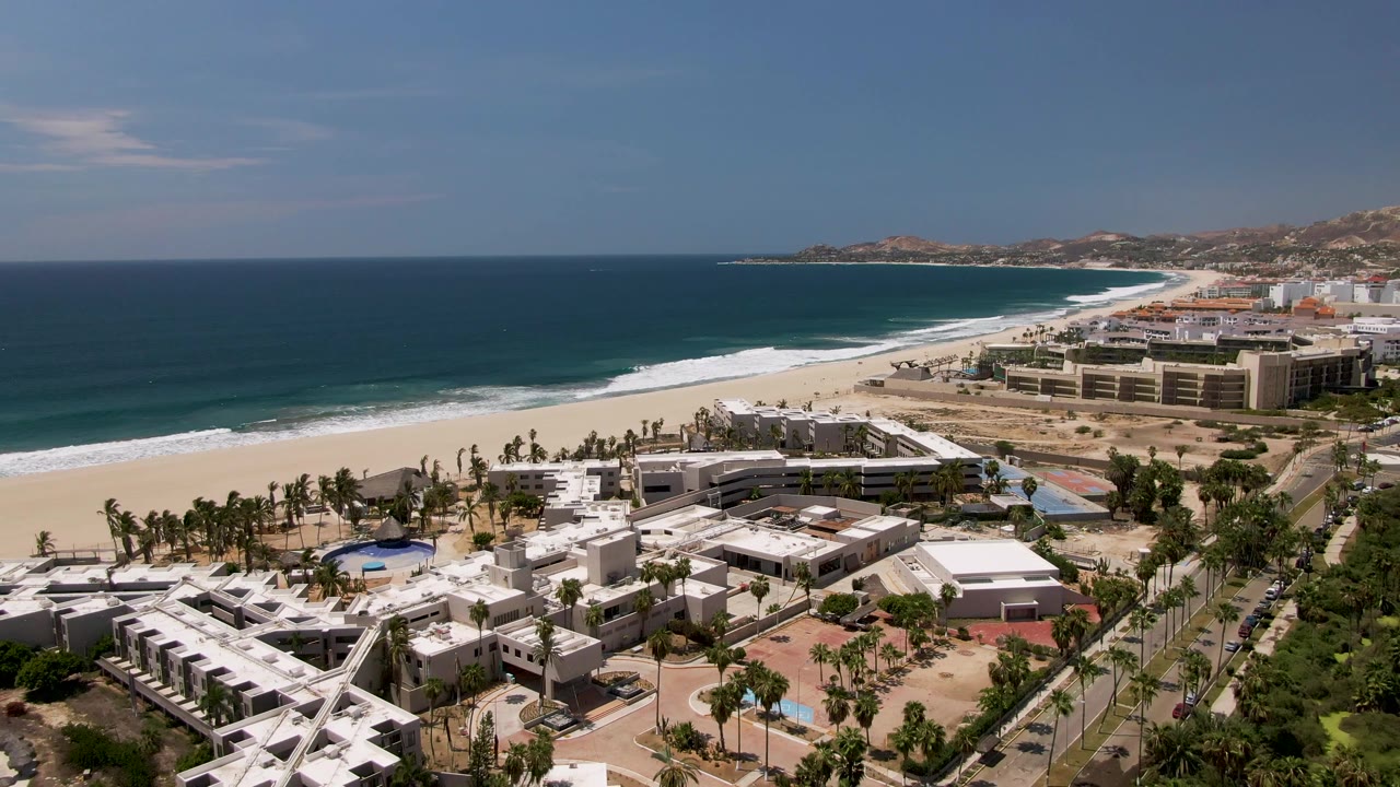 Aerial view of white resort buildings, palm trees, beach, and blue ocean.