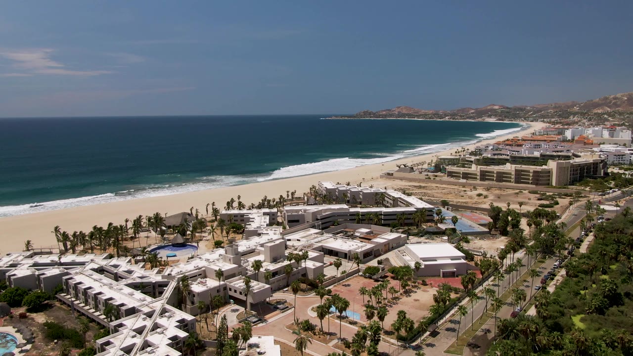 Second aerial frame in San Jose Del Cabo showing the white resort complex lower left, palms along the beach edge, a long surf line, blue water, hillside buildings in the background, and no visible people.