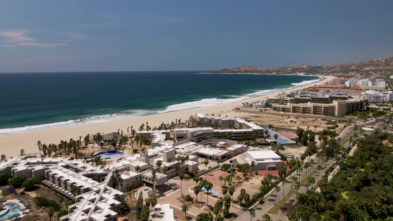 Third aerial frame over San Jose Del Cabo with the resort and road edge lower in frame, palm rows near the sand, dark blue ocean at left, bright surf along the beach, and no visible people.