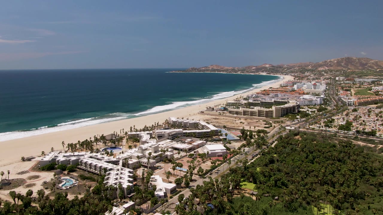 Aerial view over San Jose del Cabo with a sandy beach and blue ocean at left, resort buildings and palm trees below, hills in the distance, and no visible people.