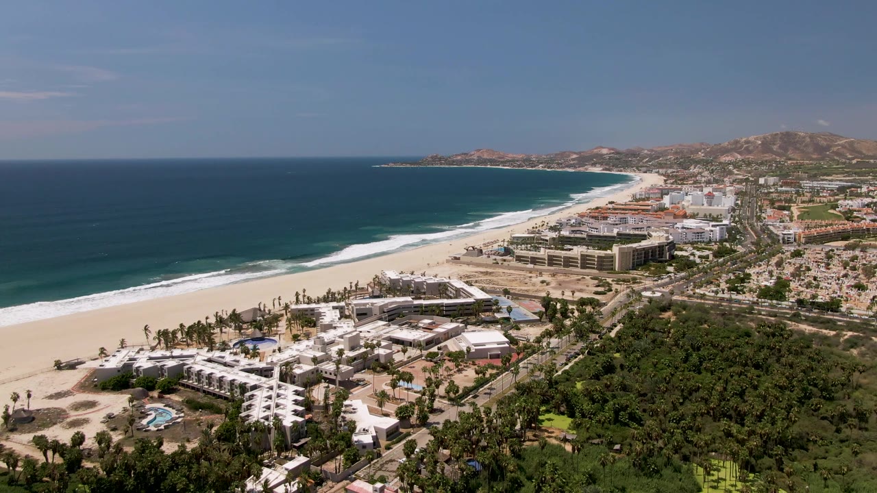 Wide aerial coastline with beach surf, resort buildings, greenery, roads, and distant hills.