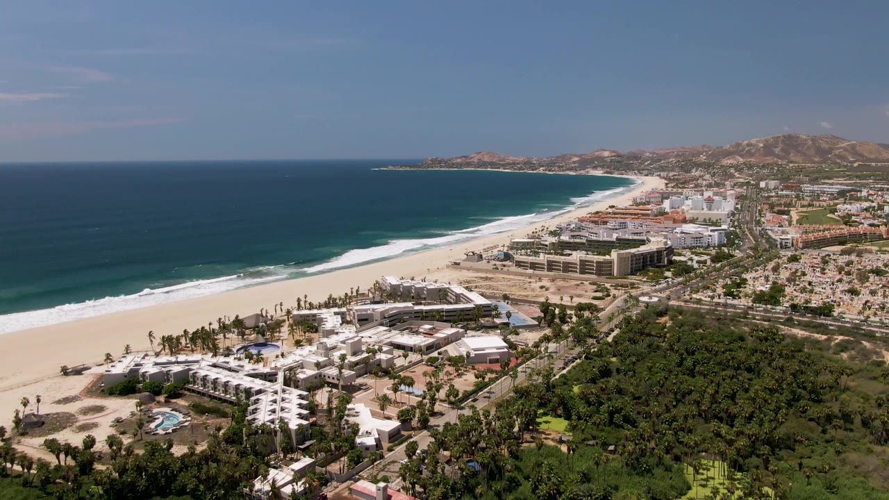 Drone-style aerial over San Jose del Cabo with the coastline curving toward distant hills, resort rooftops below, surf bands on the sand, and no visible people.