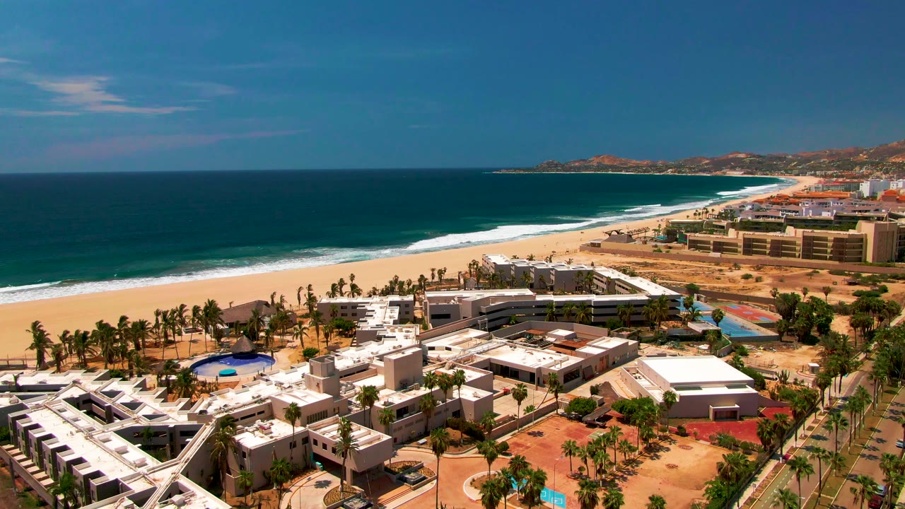 Wide aerial view in San Jose Del Cabo with resort roofs and a blue pool in the lower frame, road traffic near the beach, surf lines, distant hills, and no visible people.