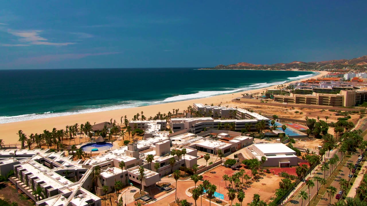 San Jose Del Cabo aerial frame with the ocean horizon high in the image, a curving sandy beach, resort blocks, palms, pools, road traffic, and no visible people.
