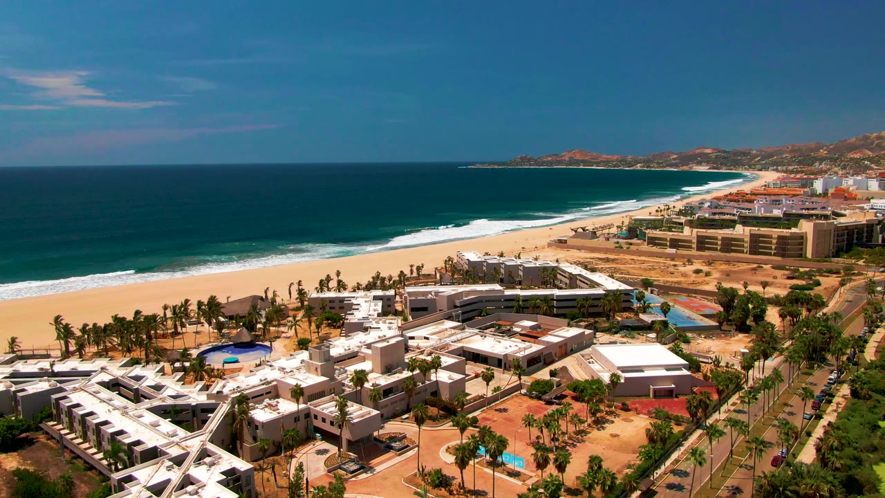 Aerial view over San Jose Del Cabo with resort buildings and palm rows below, the beach and breaking surf across the middle distance, road traffic at right, and no visible people.