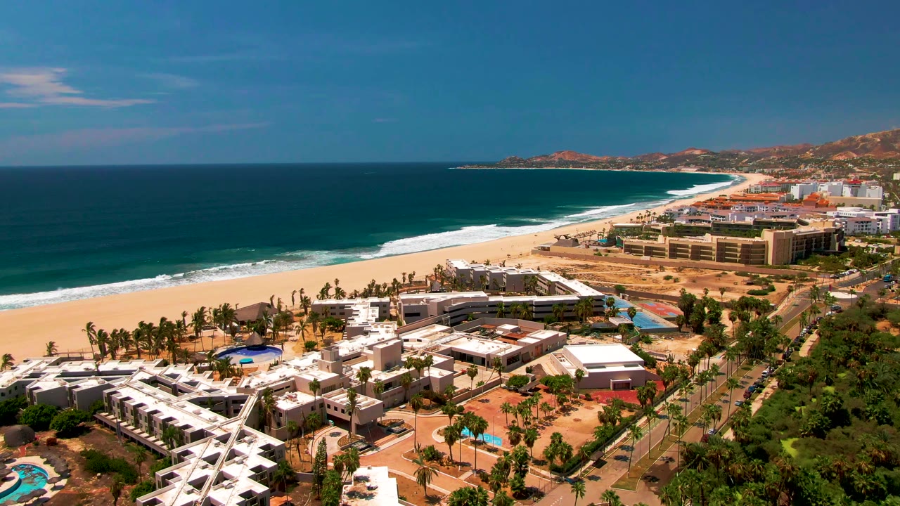 Wide elevated view of San Jose Del Cabo, Mexico, with dark blue ocean, white surf, sandy beach, resort buildings, palms, a road, green trees, and distant hills.